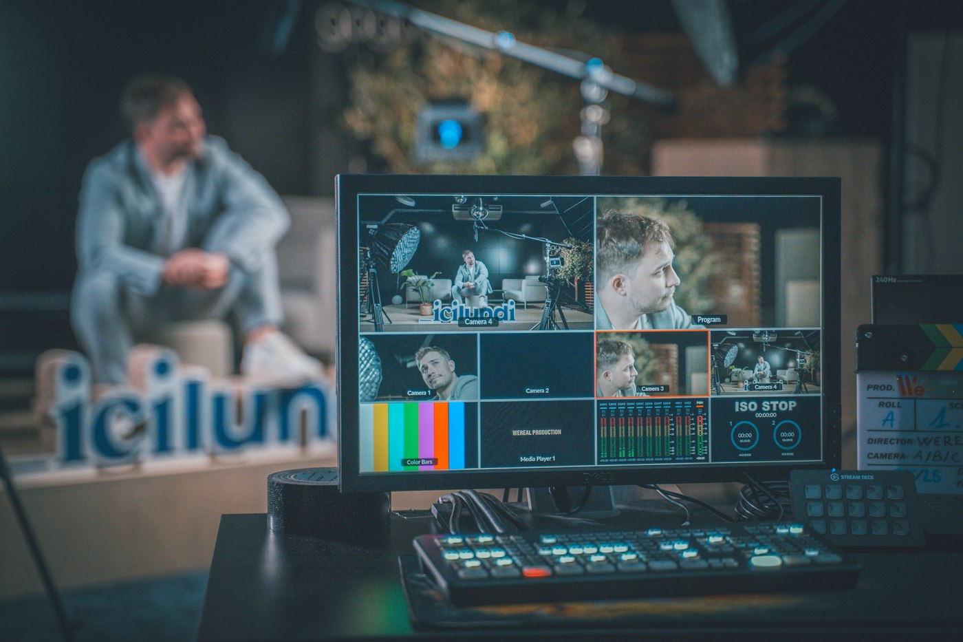 A studio control desk: a multiview monitor showing four camera feeds, with the presenter blurred behind it